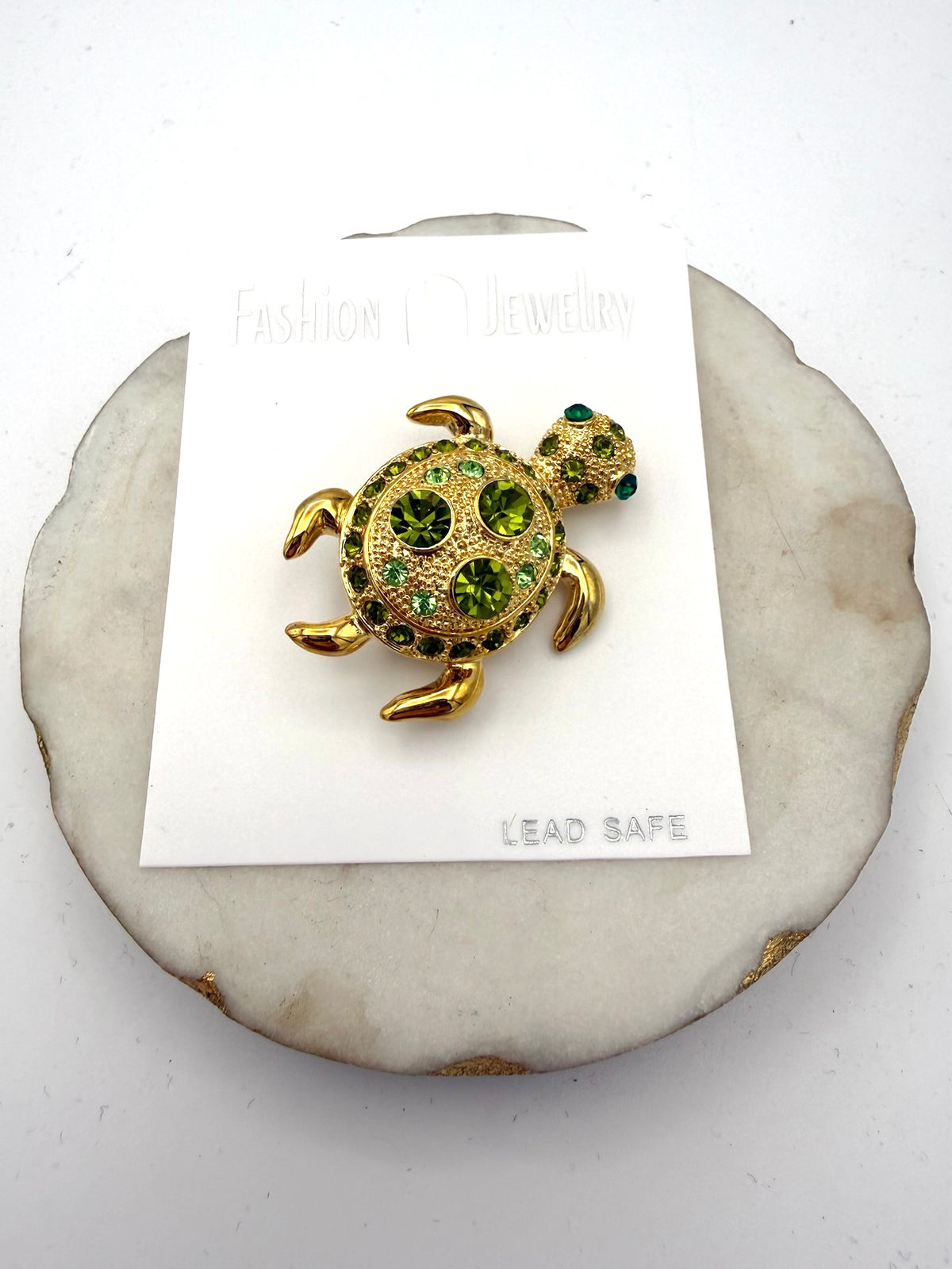 Gold-tone turtle brooch with green rhinestone shell and teal eye accent on white jewelry card