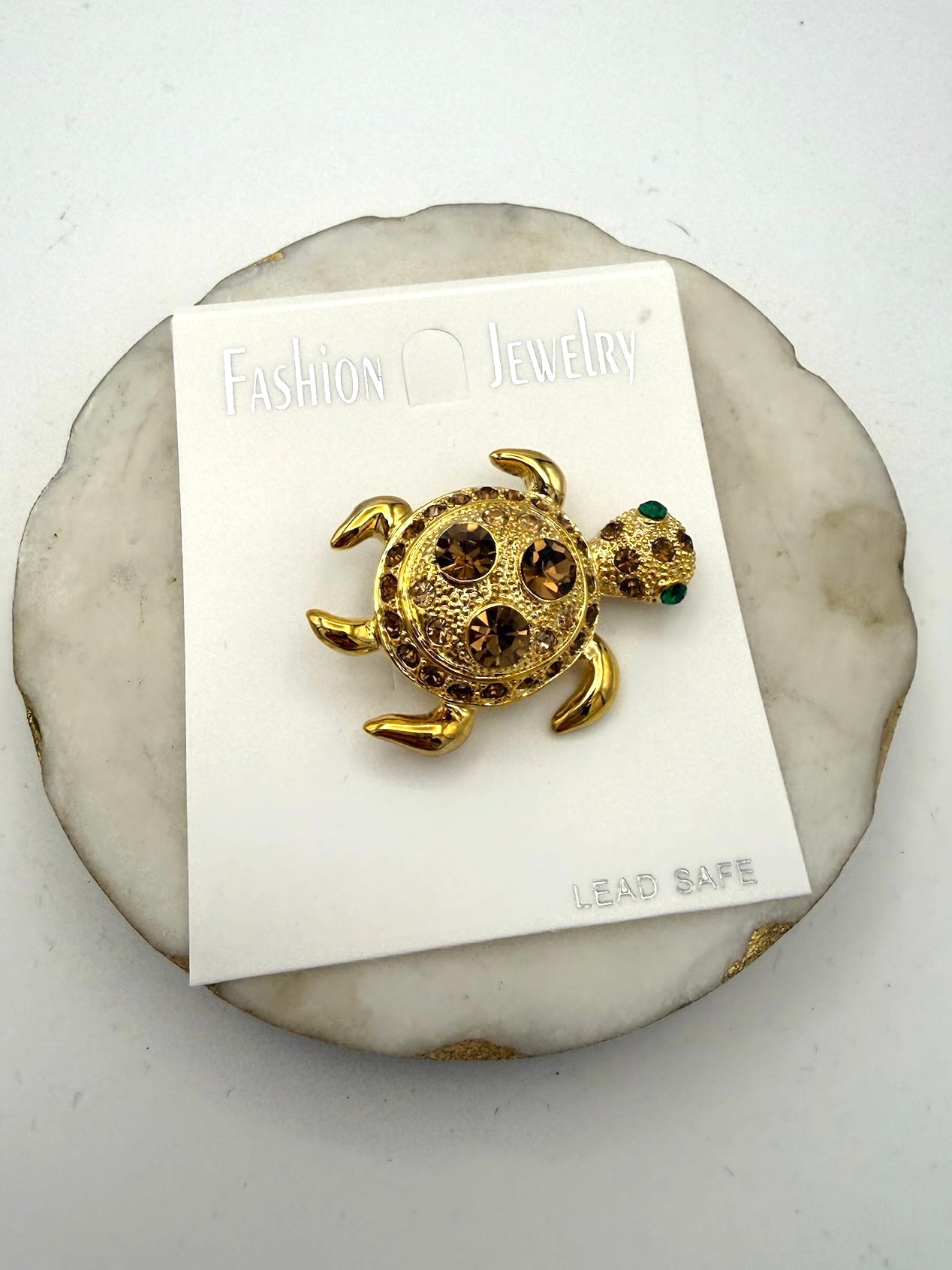 Gold-tone turtle brooch with amber rhinestone shell and green eye accent on fashion jewelry card