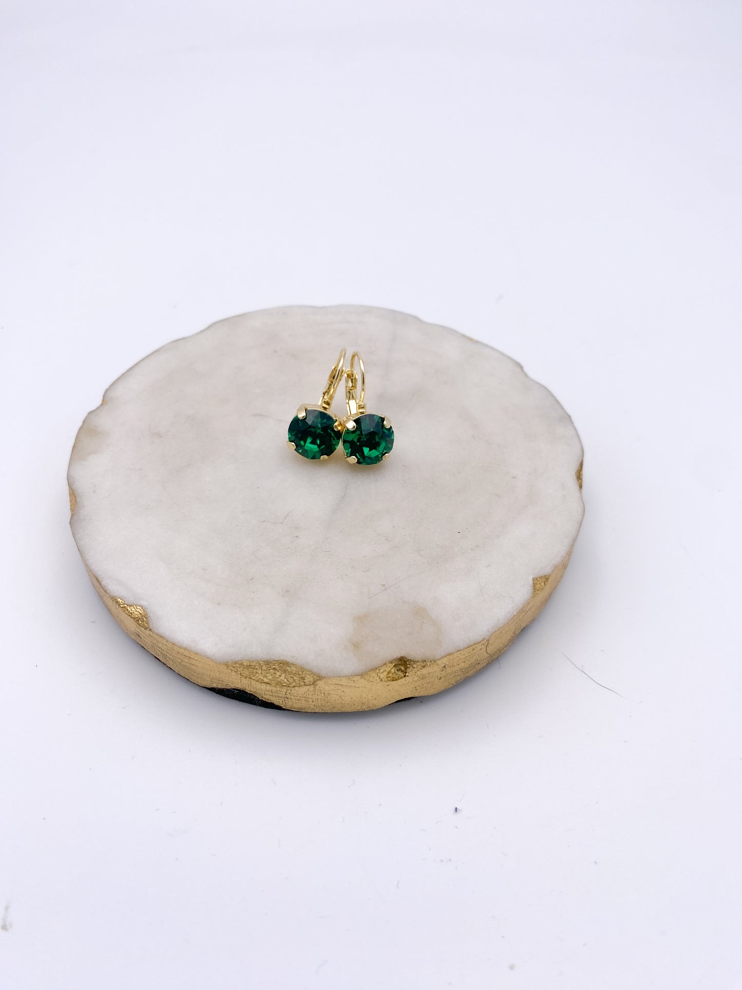 Gold lever-back drop earrings with round emerald green crystal stones, displayed on a white marble tray.