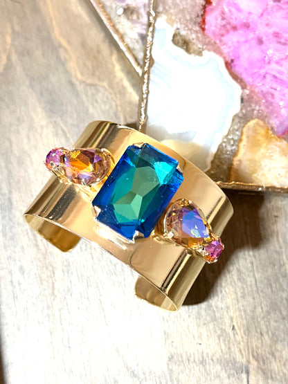 Wide gold cuff bracelet with large blue-teal emerald-cut crystal and iridescent AB accent stones.