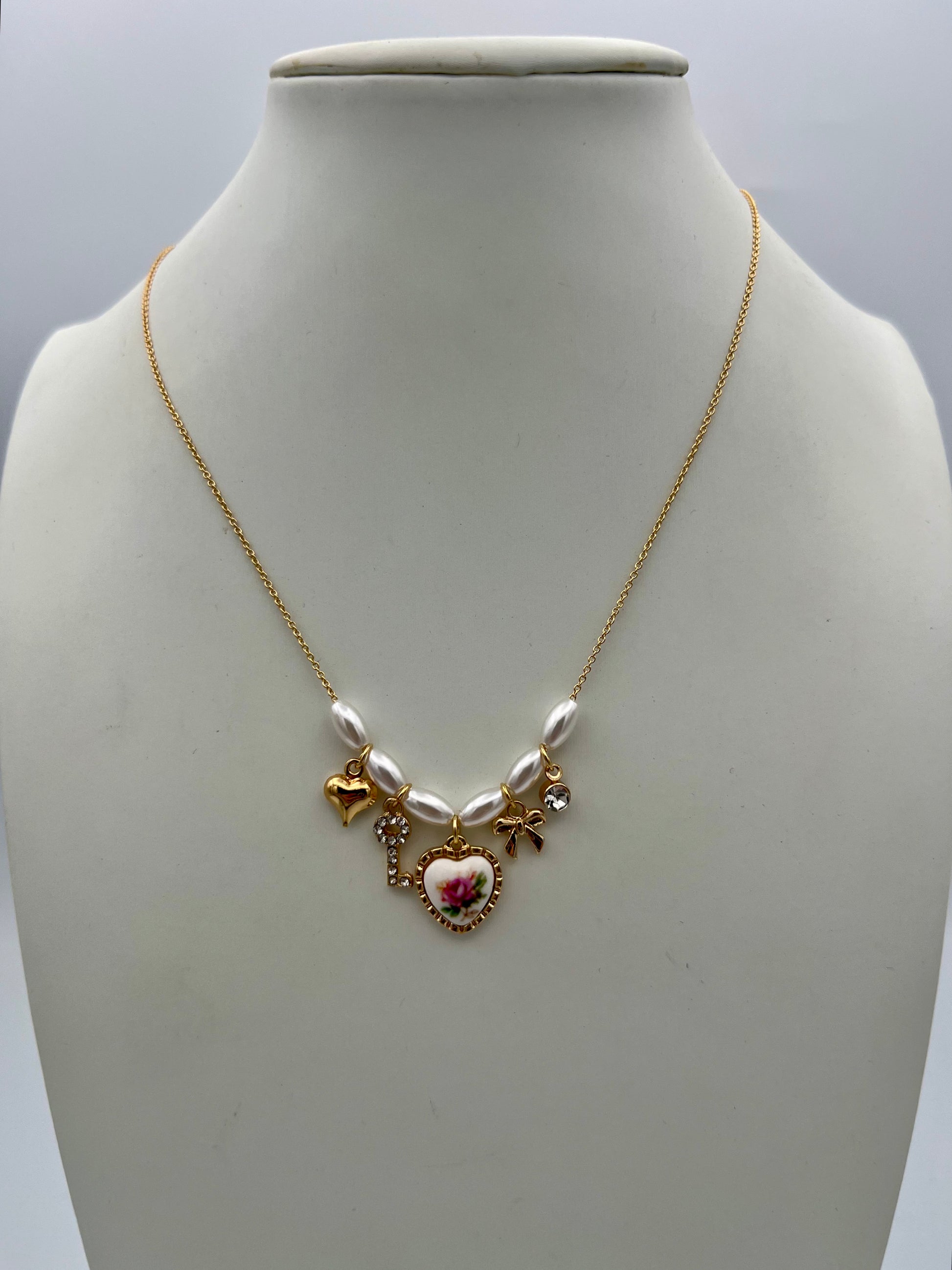 Gold chain necklace with white pearls and multiple charms including floral heart, bow, key, and rhinestone accents