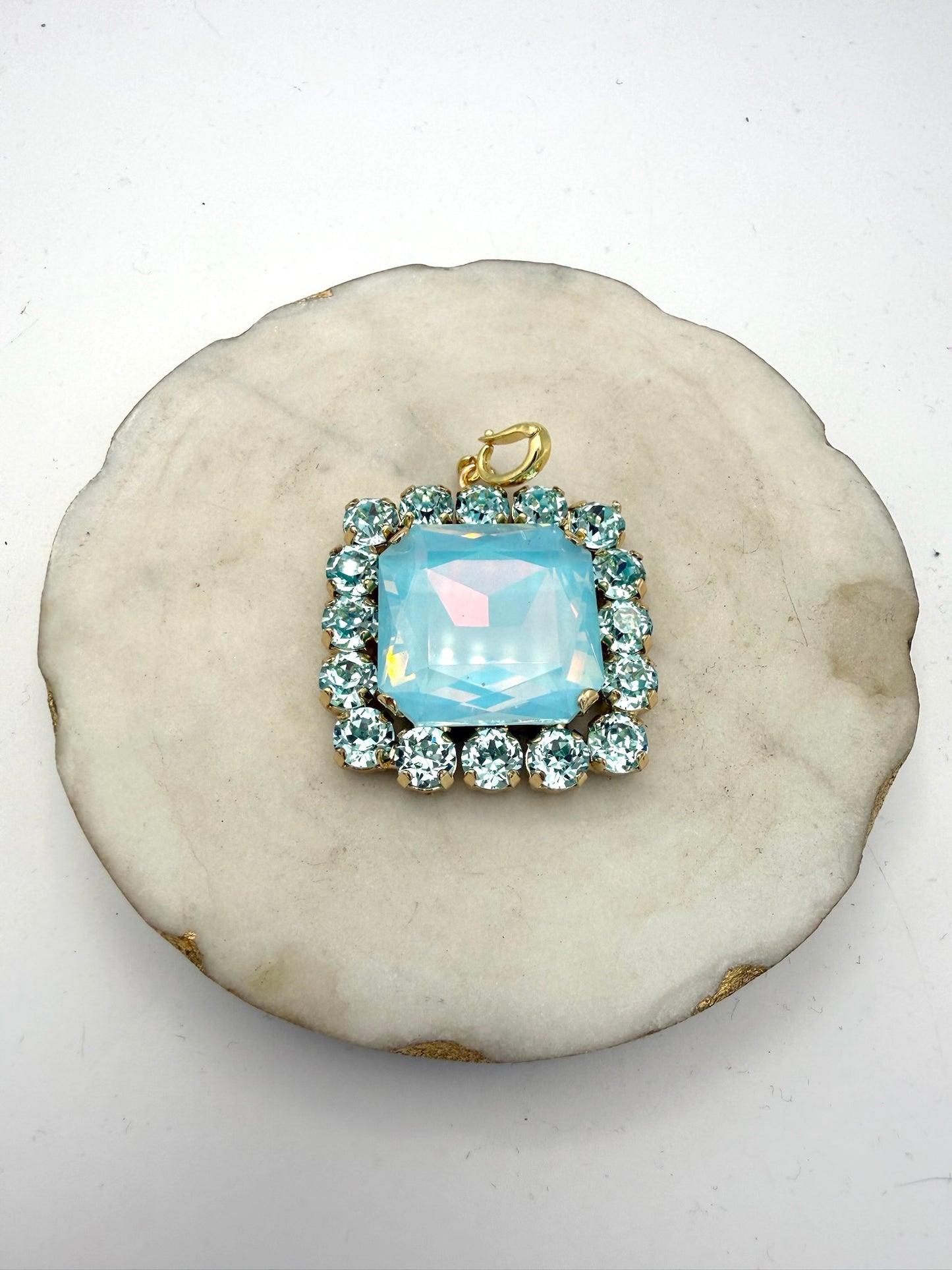 Square aqua opal crystal pendant charm with rhinestone halo border and gold bail, displayed on marble dish.