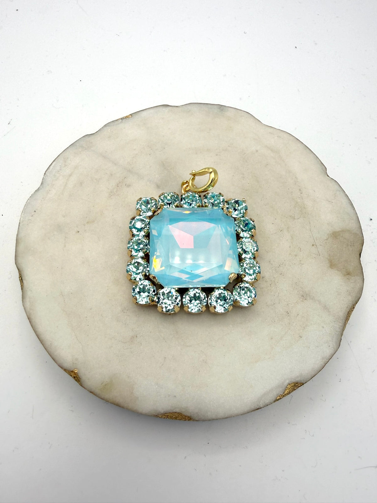 Square aqua opal crystal pendant charm with rhinestone halo border and gold bail, displayed on marble dish.