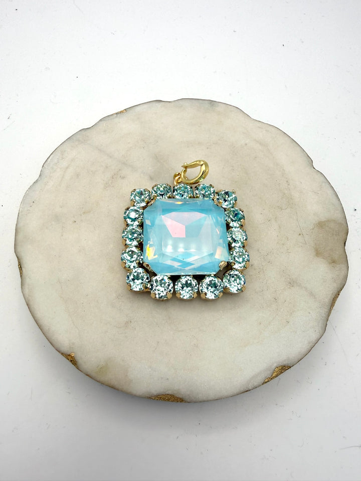 Square aqua opal crystal pendant charm with rhinestone halo border and gold bail, displayed on marble dish.
