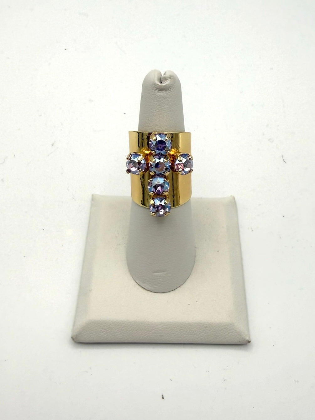 Wide gold-tone cocktail ring with iridescent lilac crystal cross motif on white jewelry display stand