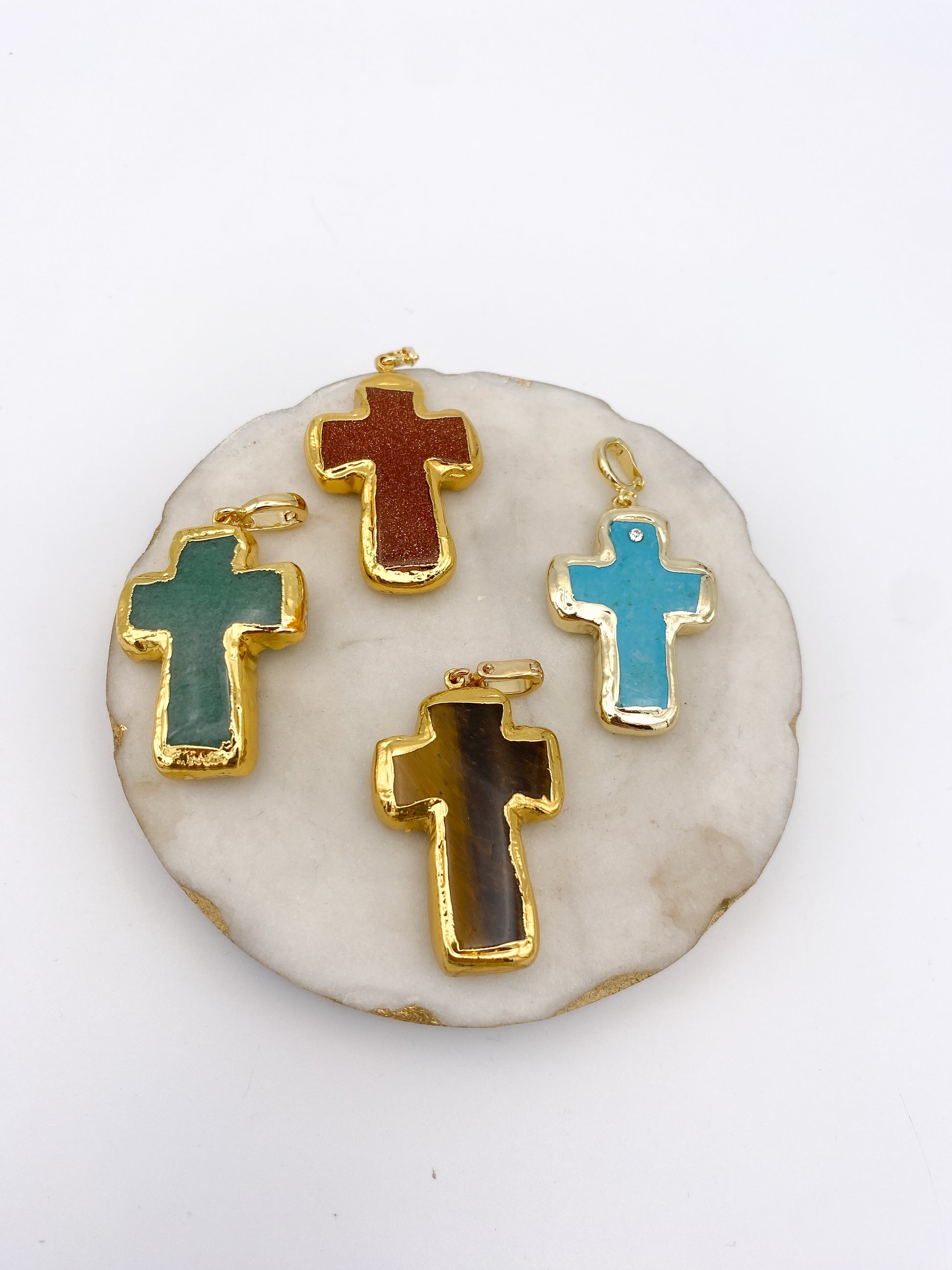 Four gemstone cross pendants with gold edging in green, goldstone, tiger's eye, and turquoise on marble display