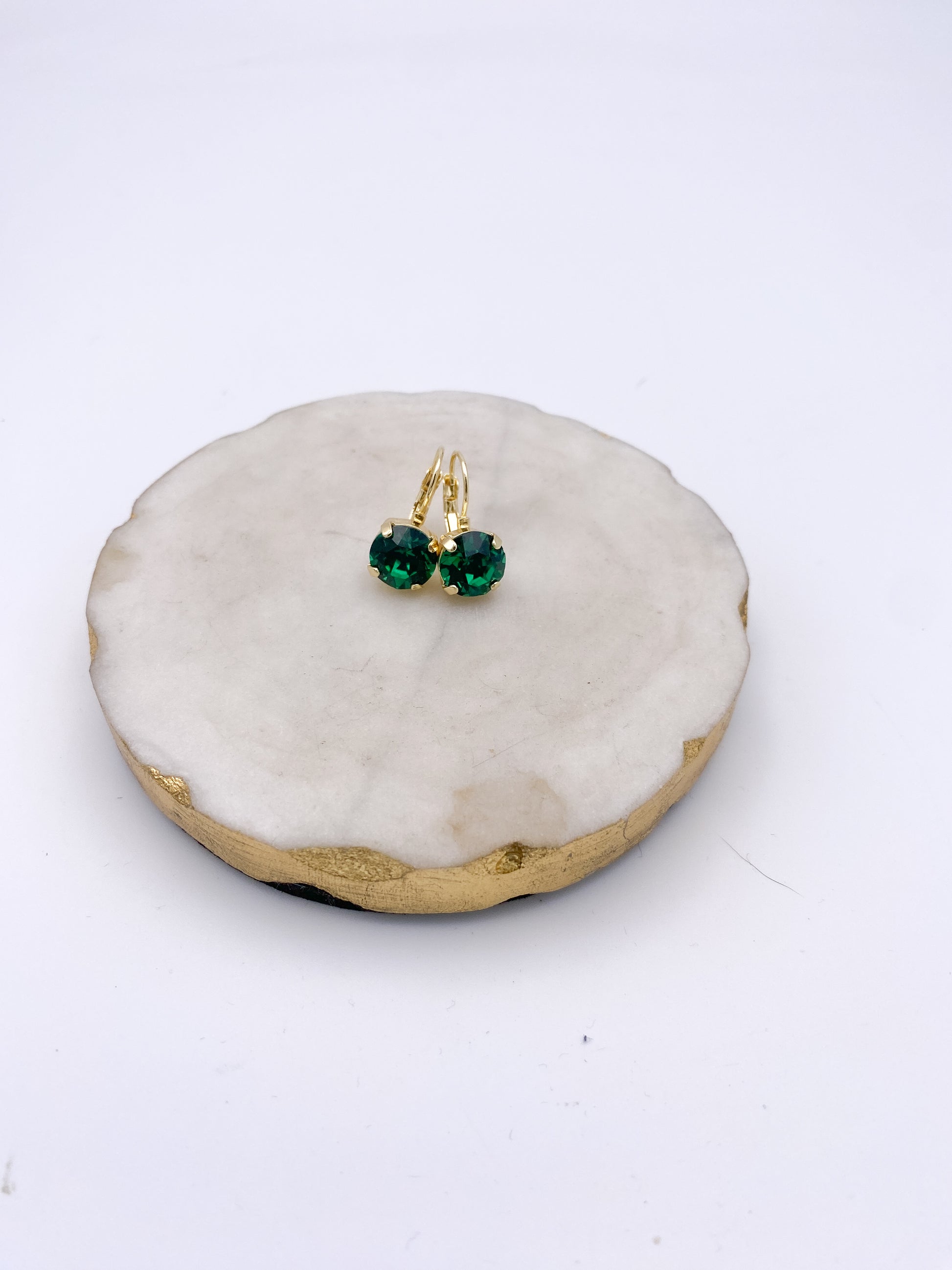 Gold lever-back drop earrings with round emerald green crystal stones, displayed on a white marble tray.